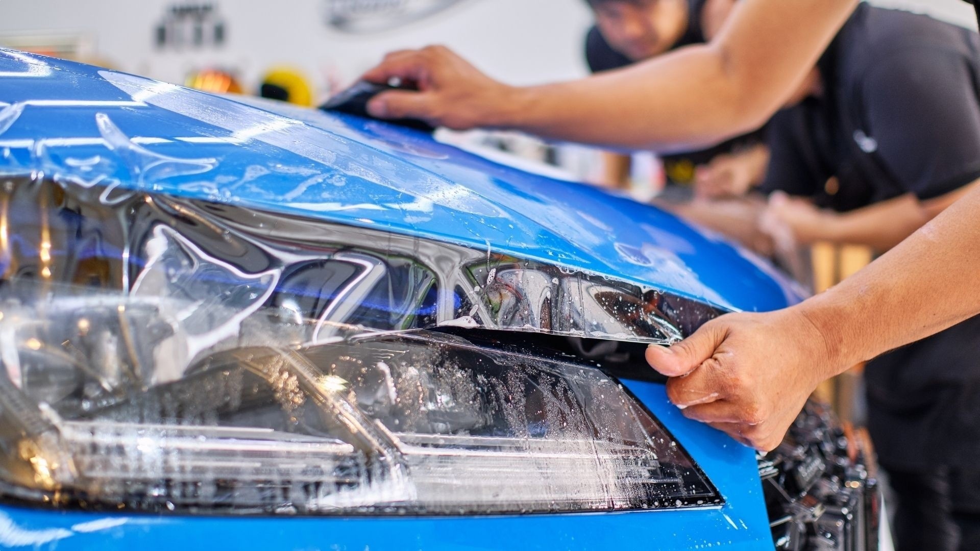 Hands smooth clear paint protection film over the front of a blue sports car.