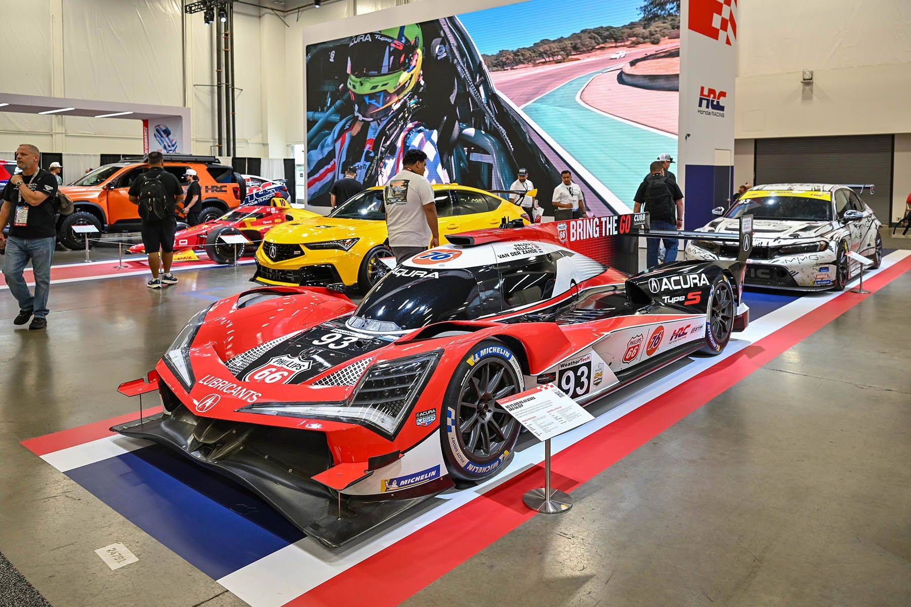 A red, black and white Acura race car displayed alongside other vehicles and visitors inside an exhibition hall.