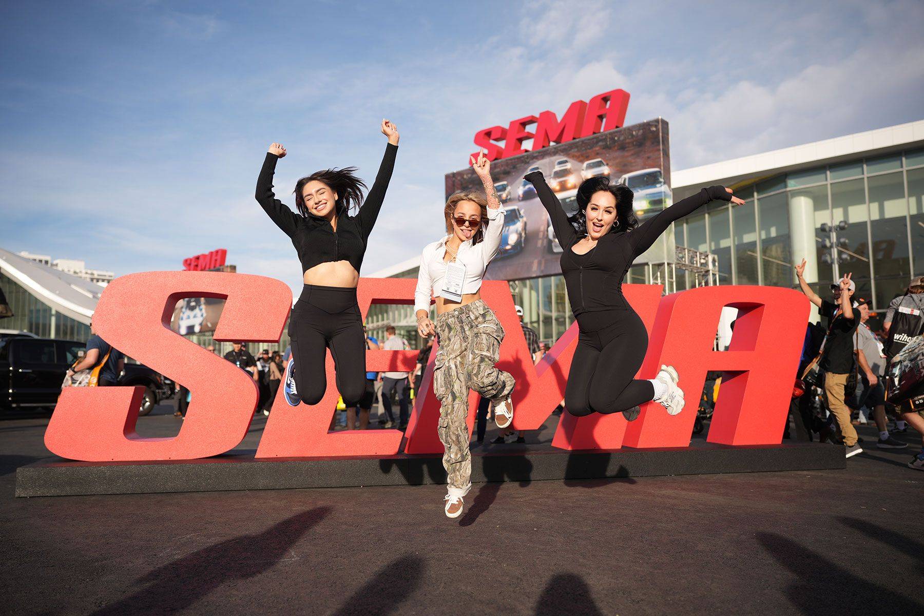 Three attendees jump in front of large red SEMA letters outside the convention center.